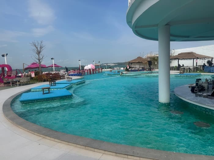 The pool at a seafront beach club, with thatched huts and the open sea beyond.