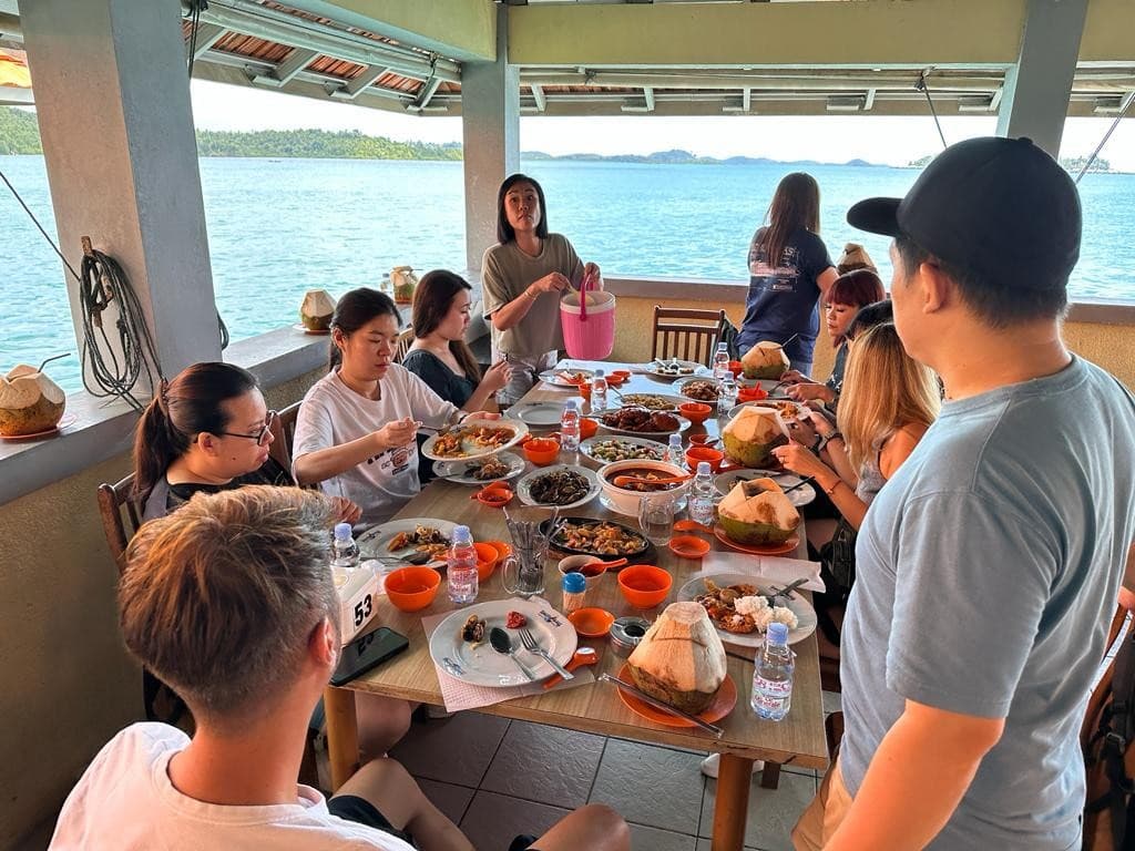 Guests having lunch at a table over the water, coconuts on the table and islands behind