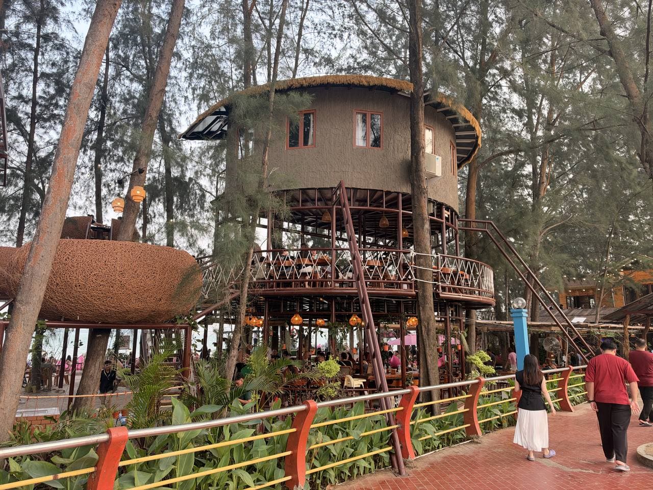 A round thatched treehouse cafe on stilts among tall pine trees, diners on the deck below
